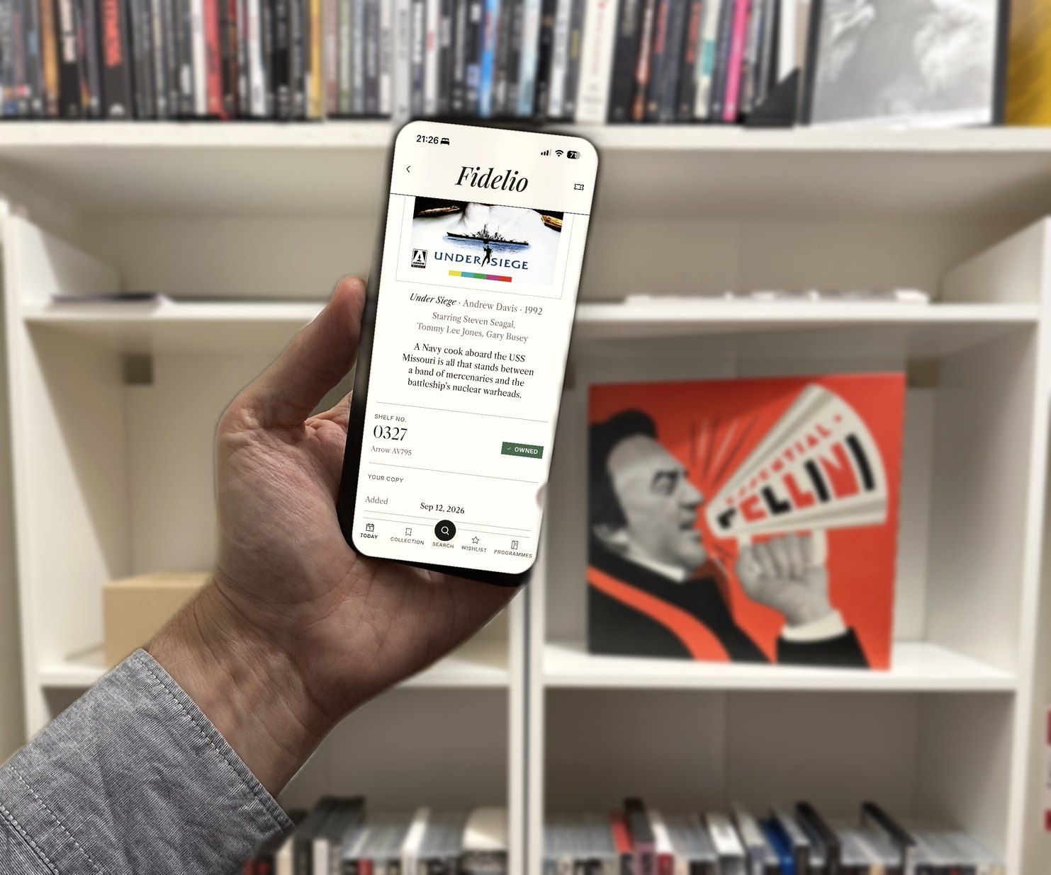 A hand holds an iPhone showing Fidelio's edition page for Arrow's Under Siege, shelf number 0327, marked owned, in front of a shelf of boutique discs and the Essential Fellini box
